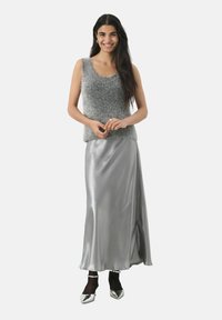 Grijze fluffy mouwloze top gecombineerd met een zilveren satijnen maxi rok. Het model draagt glanzende zilveren hakken en zwarte enkelsokken. Eenvoudig, elegant ontwerp.
