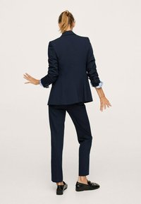 Granatowy garnitur szyty na miarę z dopasowaną marynarką i prostymi spodniami, z podwiniętymi mankietami i czarnymi loafersami, ukazany z tyłu.