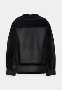 Svart shearlingjacka med plyschtextur, läderpanel på ryggen och vikta ärmslut, uppvisad mot en enkel ljus bakgrund.