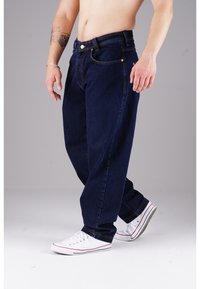 Dunkelblaue Jeans aus Denim mit lockerer Passform, sichtbaren Nähten, fünf Taschen und dekorativer Hardware. Kombiniert mit weißen Sneakers.