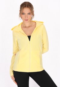 Felpa gialla realizzata in tessuto morbido, con zip sul davanti, cappuccio grande e tasche laterali. Abbinata a leggings neri per un contrasto.