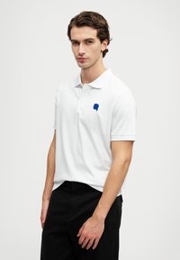 Biela polo košeľa z bavlny s modrou vyšívanou grafikou na ľavej strane hrudníka. Krátke rukávy, klasický golier, skombinovaná s čiernymi nohavicami.