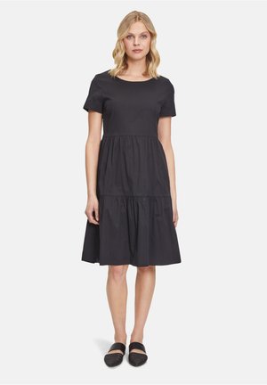 Schwarzes Baumwollkleid mit kurzen Ärmeln und rundem Ausschnitt, mit einem gestuften Rock und einem lockeren Schnitt. Getragen mit schwarzen Slippern.
