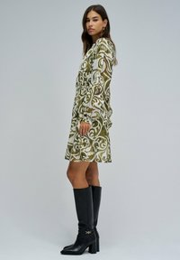 Vestido verde oliva con patrones florales blancos, de mangas largas, cintura ajustada y hasta la rodilla. Combinado con botas negras de tacón hasta la rodilla.
