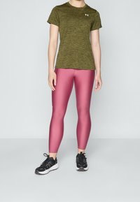 Persona con camisa de manga corta color verde oliva, leggings rosas brillantes y zapatos deportivos negros, de pie frente a un fondo liso gris claro.