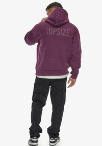 Violetinis hoodie su ryškiu „DROPSIZE“ tekstu nugaros pusėje. Derinama su juodomis krovinių kelnėmis ir sportbačiais, demonstruojant kasdišką stilių.