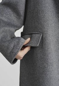 Szary wełniany płaszcz o teksturowym wykończeniu. Posiada dużą, złożoną kieszeń oraz szerokie, luźne rękawy. Wykonany z miękkiego i ciepłego materiału.