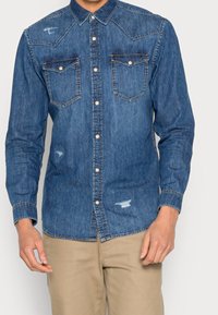 Man in een blauwe denim overhemd met knopen, voorzien van twee borstzakken en versleten details, gecombineerd met beige broek.