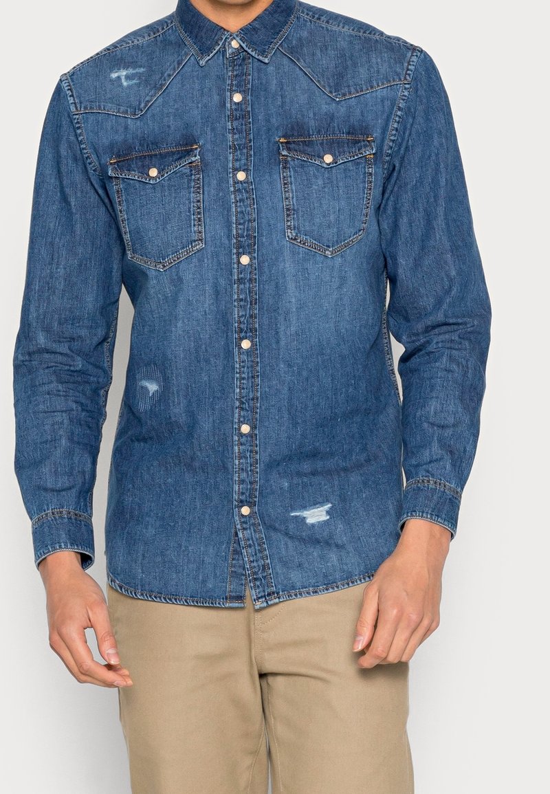 Man in een blauwe denim overhemd met knopen, voorzien van twee borstzakken en versleten details, gecombineerd met beige broek.