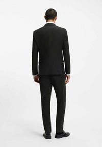 Uomo in piedi di spalle, vestito con un completo nero, le maniche della camicia bianca visibili, e scarpe nere da cerimonia su uno sfondo bianco semplice.