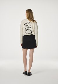 Crème lange mouwen shirt met een zwart grafisch "APÉRITIF" print op de achterkant, gecombineerd met een zwarte spijkerrok met zichtbare ritsdetail en zwarte schoenen.