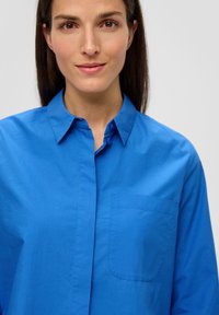Blaues Hemd mit Knöpfen aus glattem Baumwollstoff, ausgestattet mit einer Brusttasche und einem klassischen Kragen, präsentiert an einem Modell vor neutralem Hintergrund.