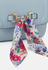 Vaaleansininen keinonahkainen käsilaukku, jossa on kultainen lukko ja kukkakuvioinen kangashuivi solmittuna kahvaan.