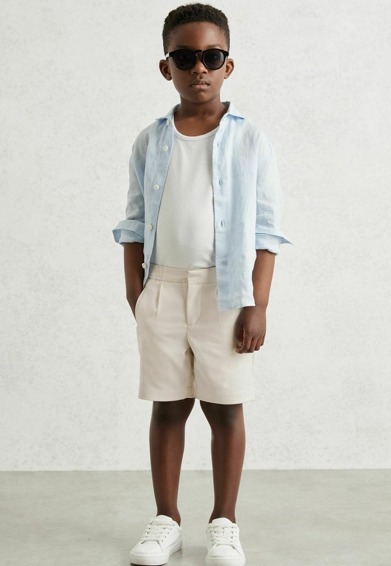 Boy wearing black sunglasses, light blue button-up shirt over white tank top, beige shorts, and white sneakers, standing with hands in pockets.