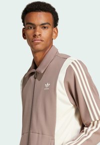 Giacca marrone con zip, colletto alto e strisce laterali bianche, realizzata in tessuto testurizzato e con logo adidas sul petto.