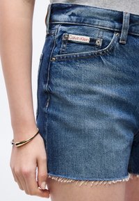 Κοντινό πλάνο ενός ατόμου που φοράει μπλε τζιν σορτς Calvin Klein με ξεφτισμένα τελειώματα και ένα χρυσό βραχιόλι στον αριστερό καρπό.