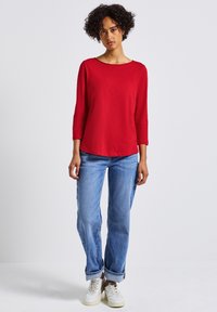 Rotes Langarmoberteil mit weitem Ausschnitt, kombiniert mit hellblauen geraden Jeans und weißen Sneakers. Glatte Textur und lässige Passform.