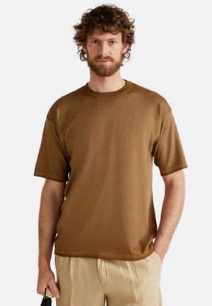 Camiseta marrón de manga corta, con cuello redondo clásico, textura suave, corte relajado, combinada con pantalones beiges claros, sostenida con una mano.