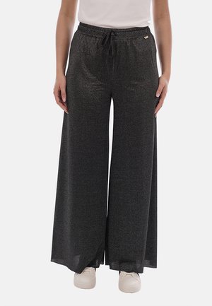 Donna che indossa pantaloni neri a gamba larga con coulisse in vita e sneakers bianche, in piedi davanti a uno sfondo chiaro e semplice.