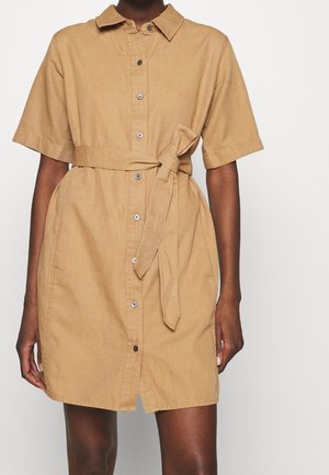Vestito camicia color beige a maniche corte con bottoni frontali e cintura annodata in vita, indossato da una persona su uno sfondo semplice.