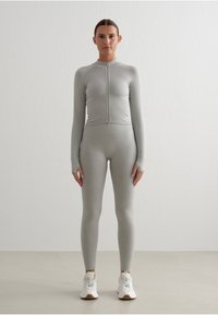 Conjunto atlético gris de corte ajustado que incluye mangas largas, un cuello alto y una cremallera frontal. Tela ligera y elástica con orificios para los pulgares.
