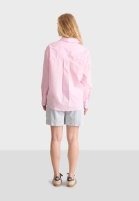 Light pink button-up shirt with long sleeves, back pleat details, paired with denim shorts. Footwear features a light-colored sole.