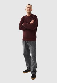 Dark red knitted sweater with a ribbed neckline, paired with faded gray jeans and black sneakers. Stands with arms crossed.