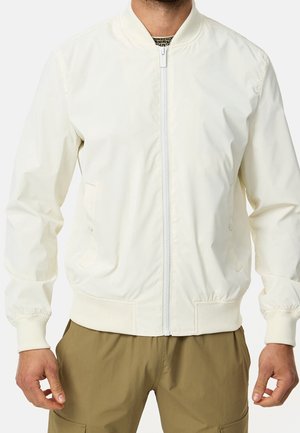 Man wearing a white zip-up bomber jacket with ribbed cuffs and hem, paired with olive green pants.