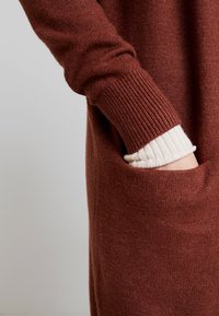 Ruskea neulottu villapaita, jossa on resorit hihansuissa; sivussa tasku ja vaaleanvärinen, resorinen pitkähihainen paidan hiha näkyy sisältä.