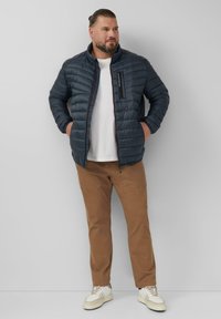 Marineblaue Steppjacke mit genoppter Textur, Frontreißverschluss und Brusttasche. Kombiniert mit hellbraunen Hosen und weißen Sneakern.