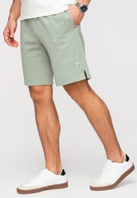 Homme portant un short vert clair avec le texte "LEVEL UP", baskets blanches et une montre, debout devant un fond uni
