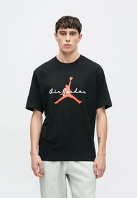 Μαύρο βαμβακερό μπλουζάκι με κοντά μανίκια που έχει ένα έντονο κόκκινο και λευκό λογότυπο "Air Jordan" και μια σιλουέτα Jumpman στο κέντρο.