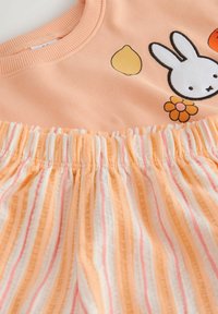 Virsiku värvi särk valge küülikuga, sidruni ja lille illustratsiooniga, mida täiendavad oranži ja valge triibulise kummipaelaga püksid.