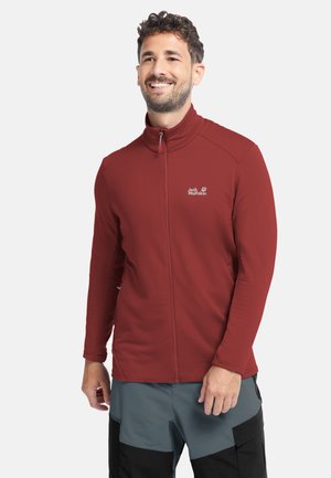 Mann lächelt, trägt eine rote Jacke mit Reißverschluss und zweifarbige graue und schwarze Hose vor einem schlichten weißen Hintergrund.