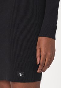 Gros plan sur une main à peau foncée reposant près d'une cuisse, portant une robe noire côtelée avec un logo Calvin Klein Jeans près de l'ourlet.