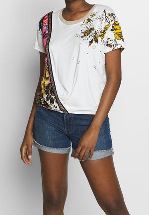 Žena v bílé košili s krátkým rukávem s žlutým květinovým potiskem a modrých džínových šortkách s ohrnutými lemy na jednobarevném pozadí.