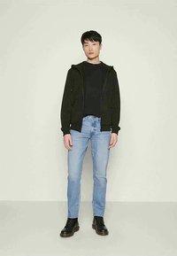 Jovem de pé contra um fundo simples, vestido com um casaco com fecho éclair preto, camisa preta, jeans azul claro e botas pretas de laço.