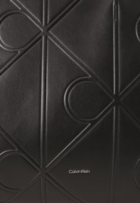 Zwarte leren tas met een geperst geometrisch patroon en de merknaam "Calvin Klein" in het wit. Glad oppervlak en strakke lijnen.