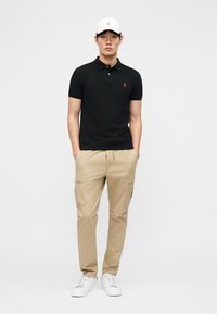 Man staat met handen in de zakken, draagt een zwarte poloshirt, beige cargobroek, witte sneakers en een witte pet tegen een effen achtergrond.