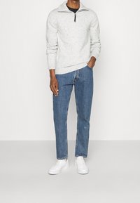 Maglione a maglia grigio chiaro con motivo a pois e collo con zip, abbinato a jeans blu a gamba dritta e sneakers bianche. Un outfit semplice e informale.