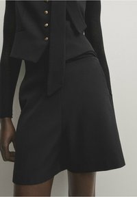 Jupe noire sur mesure avec une texture lisse, présentant une taille ajustée et un ourlet évasé. Comprend des poches latérales et des détails de design minimalistes.