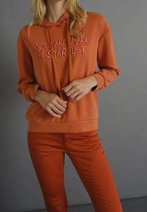 Orange hoodie with "YOU ARE MADE of STARLUST" text, featuring drawstrings and a soft texture. Paired with orange fitted pants.