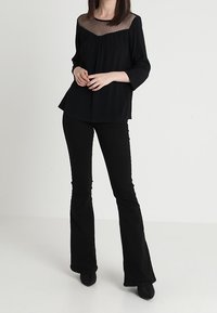 Blouse noire avec yoke en mesh transparent, motif à pois texturé, et coupe décontractée. Associée à un pantalon noir évasé et des chaussures à bouts pointus.