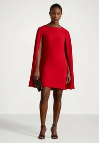Vestido mini rojo con capa, que presenta un escote redondo. Textura suave, diseño ajustado, combinado con tacones negros y un bolso de mano.