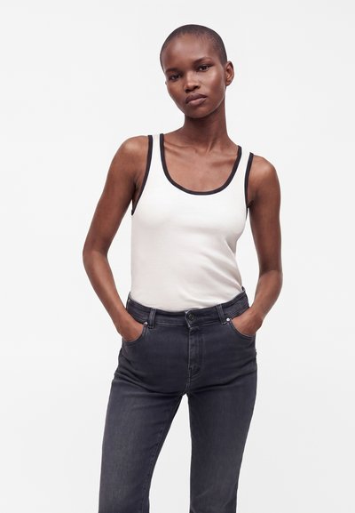 Femme aux cheveux courts portant un débardeur blanc avec une bordure noire et un jean foncé, debout, les mains dans les poches, devant un fond uni.