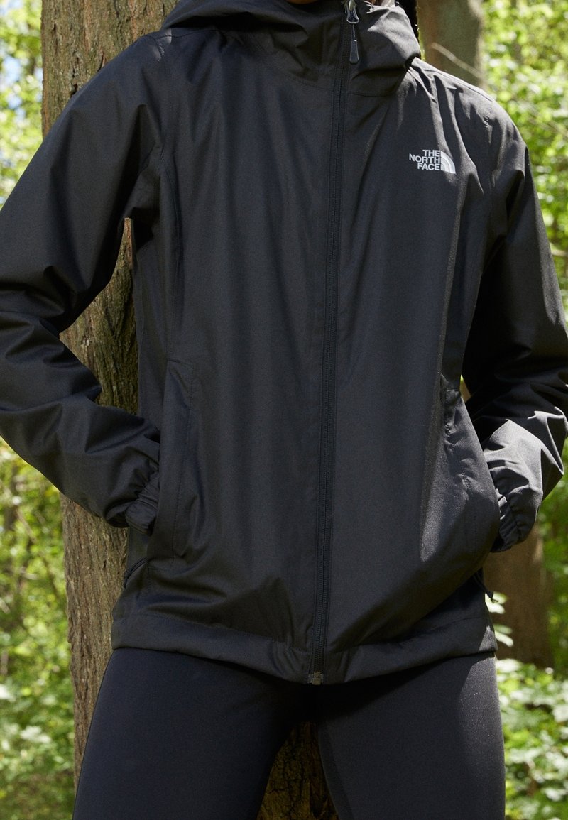 Personne portant une veste zippée noire North Face, les mains dans les poches, debout devant un arbre dans un cadre extérieur verdoyant.
