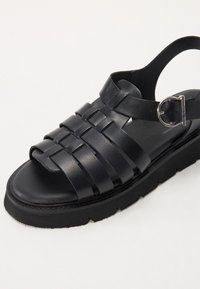 Sandale negre din piele, cu un design împletit, vârful deschis, curea la gleznă cu cataramă și o talpă groasă, texturată pentru stabilitate.