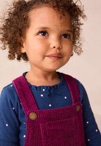 Tuta in velluto a coste viola con tasca sul petto, abbinata a una camicia a maniche lunghe blu a pois con scollatura arricciata.