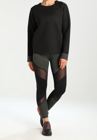 Zwarte crew-neck sweater, fitted olijfgroene leggings met zwarte meshpanelen, en zwarte sneakers met rode accenten. Glad textuur en atletisch ontwerp.