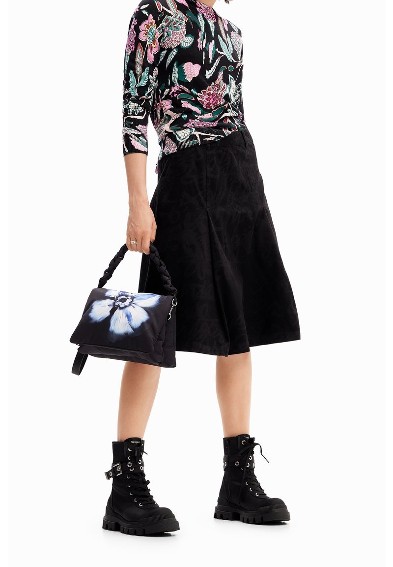Personne portant un haut à manches longues à motifs floraux, une jupe noire mi-genou, des bottes de combat noires, tenant un sac à main noir avec un grand motif de fleur bleue et blanche.